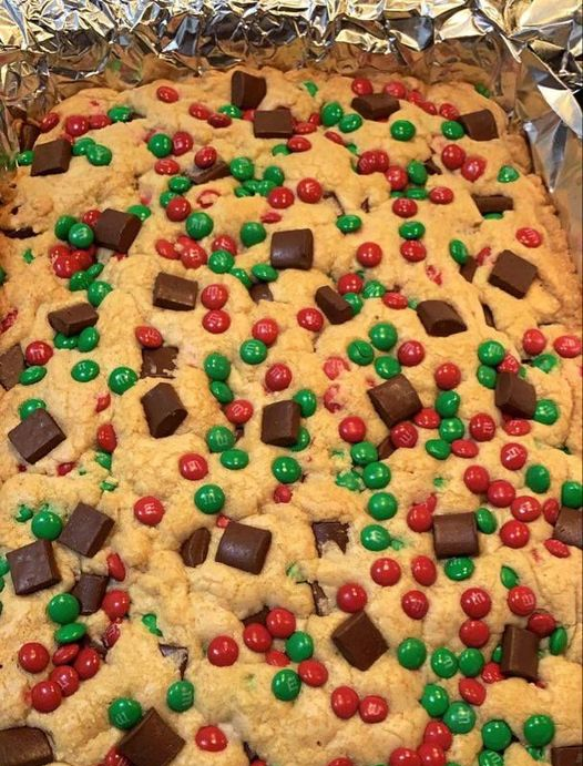 A close-up of a Christmas M&M cookie bar on a foil-covered baking sheet. The cookie bar is covered in red and green M&Ms and chocolate chips, and it has a festive holiday look.